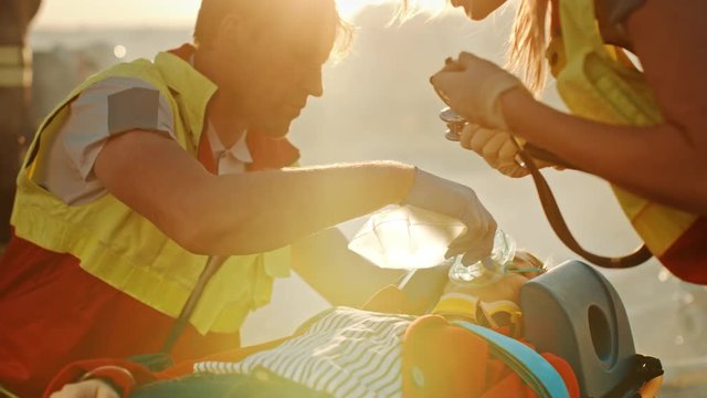 On the Car Crash Traffic Accident Scene: Paramedics Saving Life of a Female Victim who is Lying on Stretchers. They Listen To a Heartbeat, Apply Oxygen Mask and Give First Aid. Background Firefighters