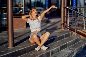 slim happy girl in shorts and white T-shirt sits on the steps of a store in the city