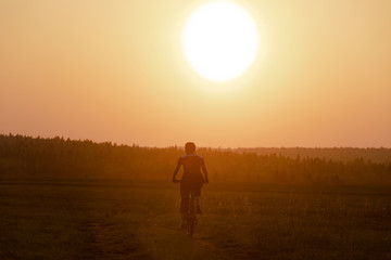 Fototapeta na wymiar A girl rides a bicycle on the road against the backdrop of a bright sunset and yellow sun