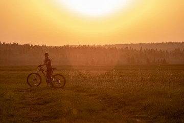 Fototapeta premium A girl rides a bicycle on the road against the backdrop of a bright sunset and yellow sun