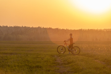 A girl rides a bicycle on the road against the backdrop of a bright sunset and yellow sun