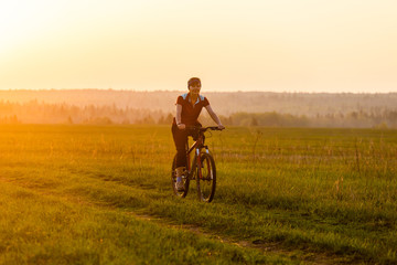 Fototapeta premium A girl rides a bicycle on the road against the backdrop of a bright sunset and yellow sun