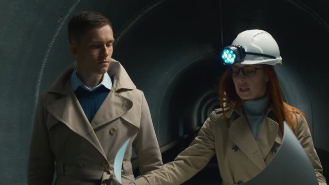 Redhead businesswoman in helmet with flashlight going through the dark tunnel with a man, showing construction plan to a man and discussing accident