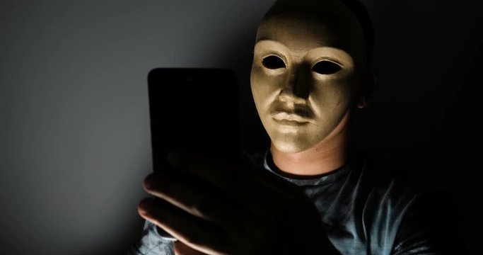 Male hacker hidden face with the mask accessing to personal information on laptop (phone) in the dark. Technology, cyber crime concept. 
