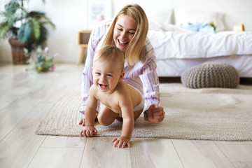 Cute cheerful European infant in diaper having joyful facial expression, laughing while crawling on...