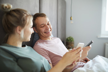 Happy guy looking at curious stuff in his girlfriend smartphone while both using gadgets in bed