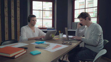 Two young man working in the office. Handsome caucasian man talking on the smartphone. guy with black hair listening music in headphones sitting at the working place using laptop. Startup team use