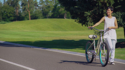 Obraz premium Adult woman with bicycle looking on beautiful nature view in the city. Elegant casual girl walking in park sunny day. Tourist with short hair wearing white shirt enjoy summer landscape.