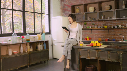 Girl drinking orange juice and reading magazine on the touch screen tablet. Attractive woman enjoy breakfast on the kitchen. Female sitting at loft room near big window at home. She using modern