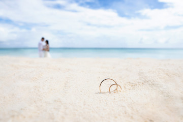 Wedding rings on the white sand. In the background the newlyweds and the ocean.