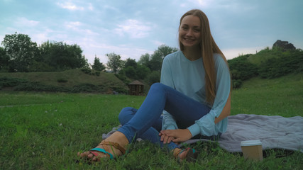 A young girl with brown hair sitting on the lawn in the park on blankets and drinking a glass of coffee. Laughing, rejoicing.