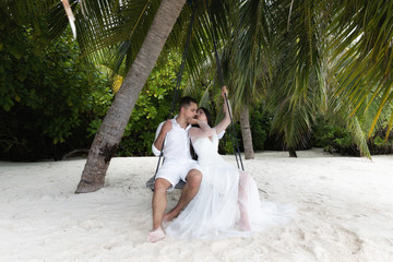 Fototapeta na wymiar Newlyweds kiss on a swing under a big palm tree.