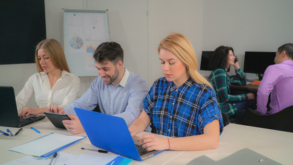 Obraz premium business team in start up company using modern device. Two blonde sitting near handsome man with beard typing on computer and talking. On the background young brunette has negotiation with client or