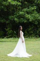 A bride in a white dress is standing on a green lawn. In the background is a tropical forest.