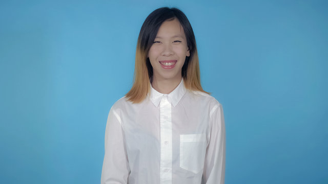 young asian happy woman posing showing positive emotions on blue background in studio. attractive millennial girl wearing white casual shirt looking at the camera smiling