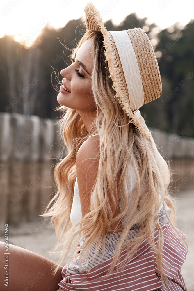 Wall mural Portrait of an attractive, romantic and c adorable provincial girl with long loose hair in a straw hat. Sunset, soft sunny colors. Nature landscape. Countryside at the background. Summer, summertime