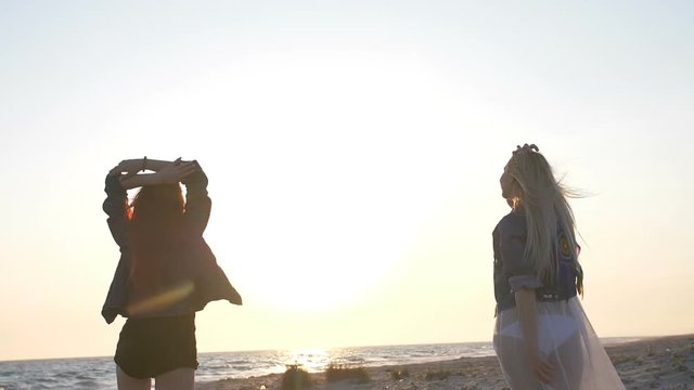 dance on the beach of two young sexy women dressed in boho style in the sunset light over the sea
