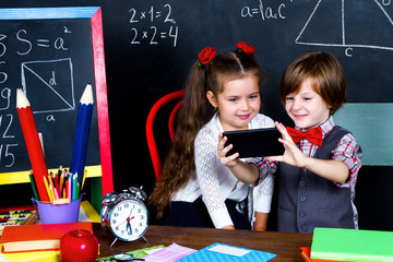 Intelligent smart happy schoolboy and schoolgirl playing in mathematic teacher opposite school chalkboard at school in classroom with school books, notebooks and smartphone. Back to school concept