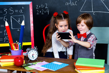 Intelligent smart happy schoolboy and schoolgirl playing in mathematic teacher opposite school chalkboard at school in classroom with school books, notebooks and smartphone. Back to school concept