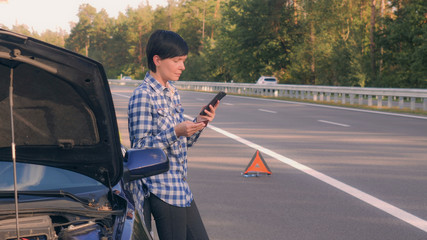 Adult brunette standing near broken car holding card entering phone number on smartphone calling evacuator or customer service. Lady standing on the road using mobile phone in day time summer season.