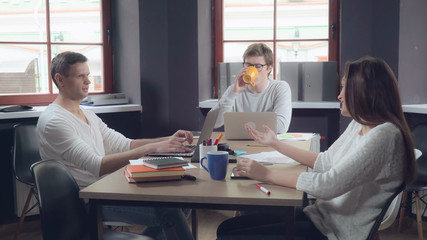 Obraz premium Brainstorming in the contemporary office. Young professional people talking using computer paper documents dressed in casual clothes. Two handsome man and caucasian woman discussing project.