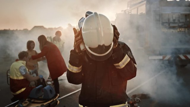 Portrait of the Brave Firefighter Taking Off His Helmet. In the Background Courageous Heroes Paramedics and Firemen Rescue Team Fight Fire, Smoke and Save People&rsquo;s Lives