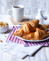 Delicious breakfast french style at home: light background, white plate with fresh croissants, small bowl of granola, cup of tea and bottle with milk. Served on red and beige napkins