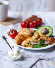 Delicious breakfast french style at home: light background, white plate with fresh croissants, small bowls of granola and cream cheese, cup of tea, avocado, tomatoes, basil. Served on beige napkins