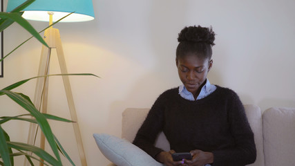 Woman messaging sms on smartphone sitting on the cozy sofa in apartment. Beautiful lady chatting online with happy smile on her young face. Afro american model surfing internet on cell phone.