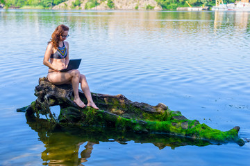 Freelancer working on a laptop while sitting on a fallen tree near the river.
