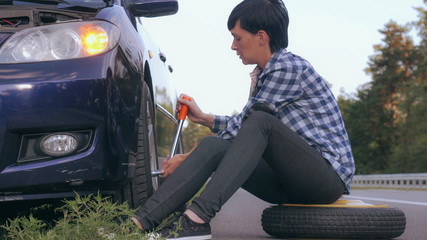 Young woman stuck on the side of the road with a flat tire. Caucasian brunette driver change a tire at the roadside.