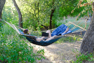 Business woman working on laptop while on vacation. Young woman lying in a hammock and looking to a laptop.