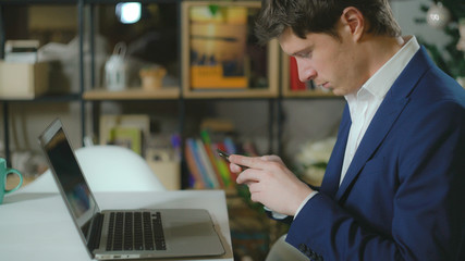 Handsome man sitting in room with bookshelves on the desk laptop. Elegant fashionable man wearing in casual blue suit texting message on smartphone.