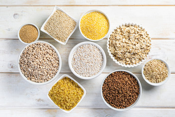 Selection of whole grains in white bowls - rice, oats, buckwheat, bulgur, porridge, barley, quinoa, amaranth, on white wood background