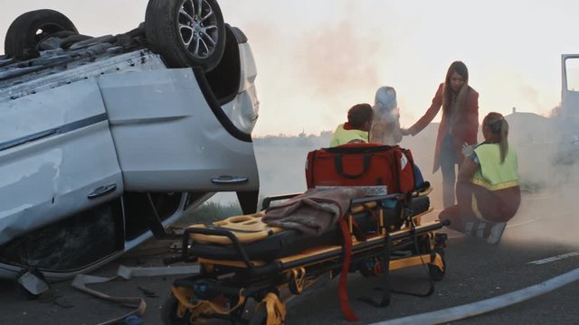 Burning Rollover Car in a Crash Traffic Accident: Team of Paramedics Perform Emergency First Aid to Injured Mother and Young Daughter. Giving Oxygen Mask and Checking for Trauma