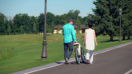 Friends walking with bike rear back view. Woman and man going in park in sunny day. People wearing casual dress active leisure at summertime outdoors.