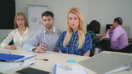 portrait three co workers sitting at the table posing looking at the camera. On the background staff talking with happy smile.