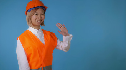 portrait young asian female posing wearing uniform orange hard hat and vest waving hand say hello on blue background in studio. attractive korean woman with blond hair wearing white casual shirt