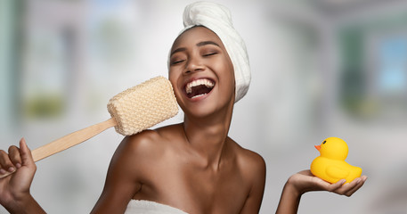 Close-up portrait of a beautiful smiling happy woman holding body scrubber and yellow rubber duck isolated on bright background. Beauty treatments and cosmetics spa therapy