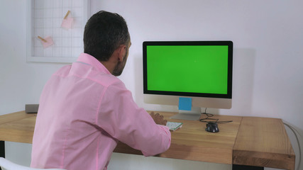 Back view focused businessman wearing casual shirt sitting at the working place works on computer with green display. Man entering data typing on keyboard in home office looking on pc