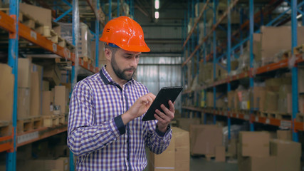 Young manager working in warehouse. Handsome worker using touch screen tablet check stock entering information. Man wearing casual shirt and hard hat at work.