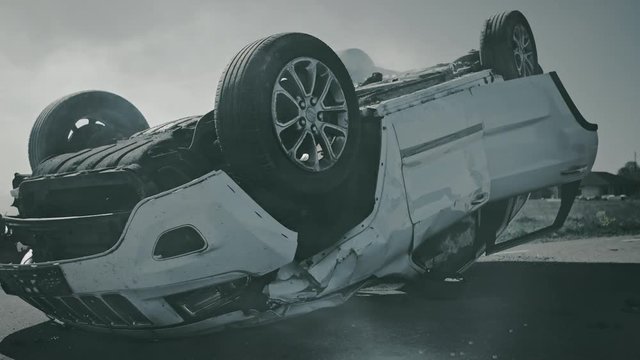 Horrific Traffic Accident Rollover Smoking and Burning Vehicle Lying on its Roof in the Middle of the Road after Collision. Daytime Crash Scene with Damaged Car. Arc Shot In Black and White