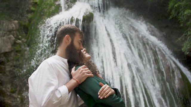a man with a beard embraces a woman with curly hair behind her who leaned on his chest against the backdrop of a waterfall in a mountain forest
