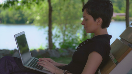 Girl with short haircut and black hair using laptop outdoor. Young woman sitting on the bench in park with lake typing on computer. Lady wearing casual polo shirt working on pc in summer season.