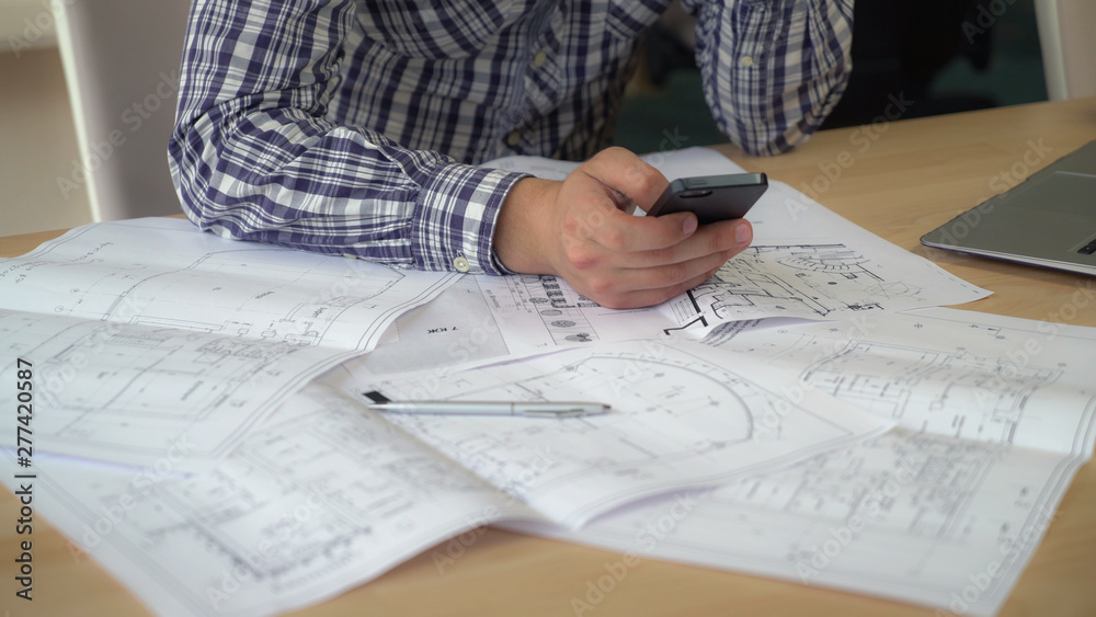 Aufkleber Designer using mobile phone works with plan building. take photo of sketch and send client or sharing in social net. Man hands holding smartphone texting message at the working place. On the wooden