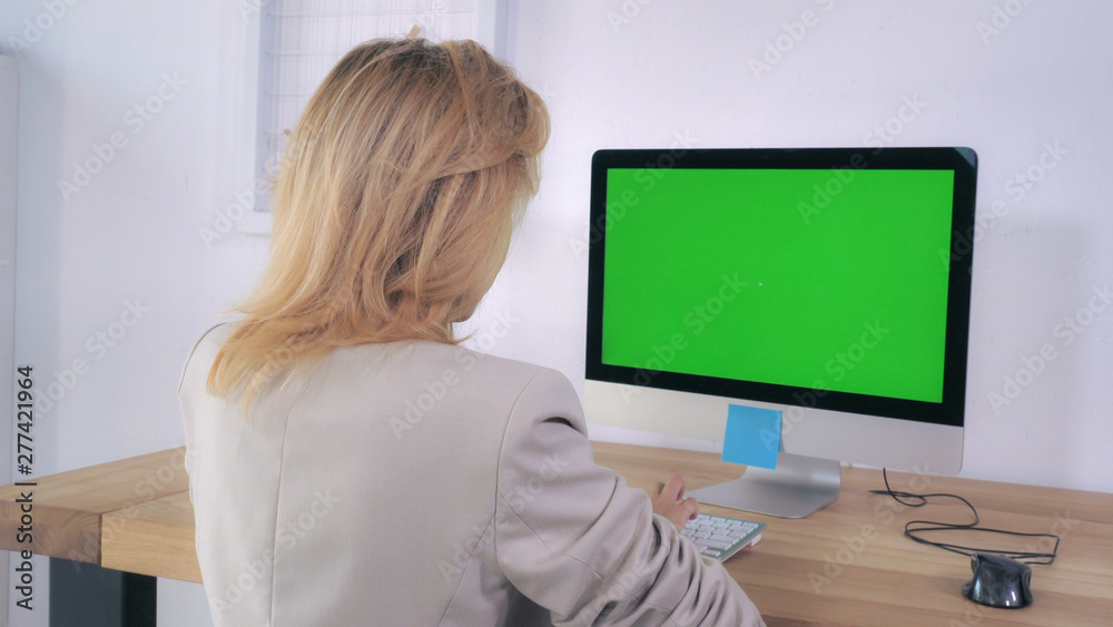 Poster Blonde typing keyboard sitting at the working place with computer on the wooden desk display with green screen. Back view woman using pc in room with white wall