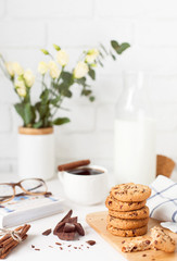 Morning breakfast with a cup of coffee with cinnamon, tasty cookies with chocolate, a bottle of milk, a bouquet of flowers and a book on the background of a white brick wall