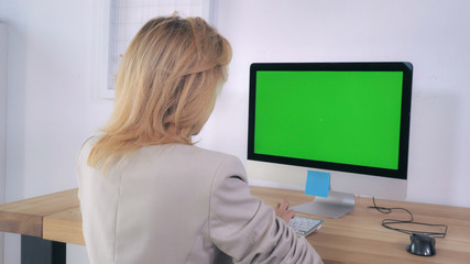 Fototapeta na wymiar Blonde typing keyboard sitting at the working place with computer on the wooden desk display with green screen. Back view woman using pc in room with white wall