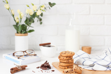 Morning breakfast. A cup of coffee with cinnamon, delicious cookies with chocolate, a bottle of fresh milk, a bouquet of flowers and a book with glasses on a white brick wall background 