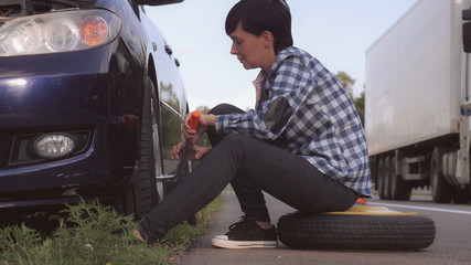 Caucasian young woman wearing casual shirt and jeans changing a tire sitting on stepney near car on the roadside. Brunette traveller outdoors